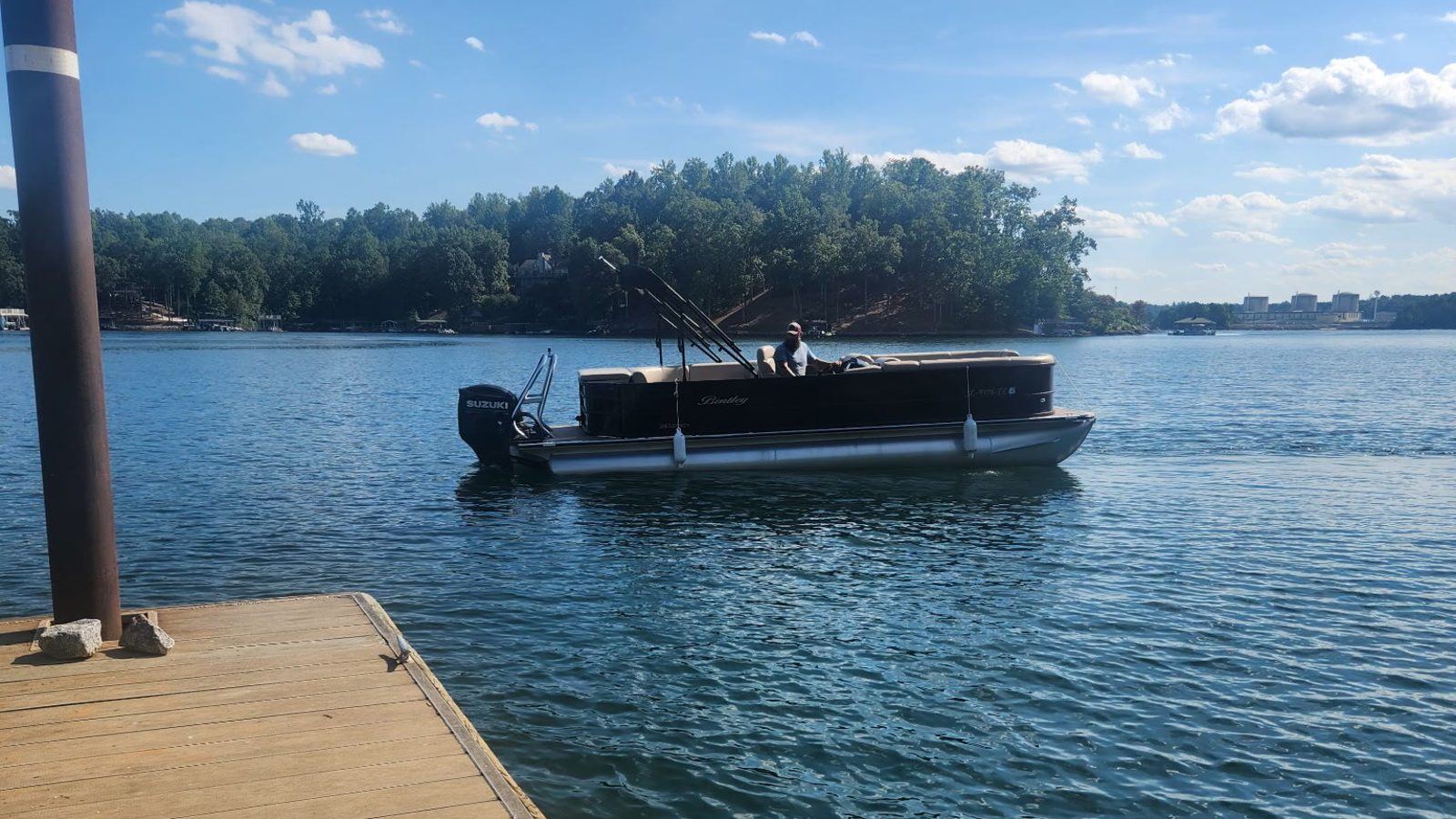 A black tritoon cruising across open blue water on Lake Keowee, seen from the end of a wooden dock, with a wooded point behind it.